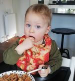 Das Bild zeigt ein männliches Baby, das gerade Nudeln isst. 