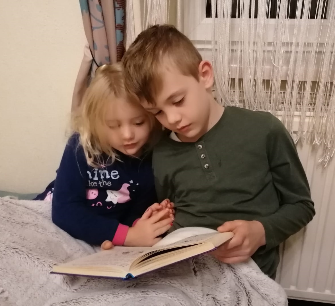1 Mädchen (5 Jahre) und 1 Junge (ca. 8 Jahre) sitzen zusammen im Bett. Der Junge hält ein Buch in der Hand und es sieht aus, als würde er dem Mädchen vorlesen