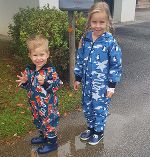 Ein 5-jähriges Mädchen und ein 2-jähriger Bub tragen bunte Overalls, Gummistiefel und stehen im Regen. Ein 5-jähriges Mädchen und ein 2-jähriger Bub tragen bunte Overalls, Gummistiefel und stehen im Regen.