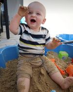Ein ca. 1-jähriges Kind sitzt in einer Sandkiste im Garten. Neben ihm sind Sandspielsachen zu sehen und er trägt ein gestreiftes Shirt.