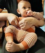 Das Bild zeigt ein männliches Baby, das auf einem Schoß gehalten wird. Das Baby trägt einen gestreiften Strampelanzug.