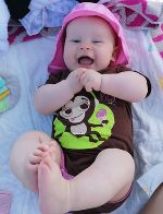 Das Bild zeigt ein vergnügtes weibliches Baby mit Kappe in einer Wippe liegend und lachend. Es ist barfuß.