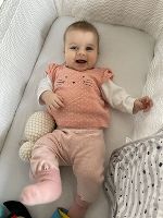 Ein Säugling (Mädchen) liegt im Gitterbett und schaut vergnügt in die Kamera.