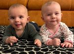 Das Foto zeigt zwei Babys, die nebeneinander auf einer gemusterten Decke liegen und in die Kamera schauen.