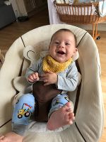 Das Bild zeigt ein vergnügtes männliches Baby mit braunen Haaren in einer Wippe liegend und lachend. Es ist barfuß.